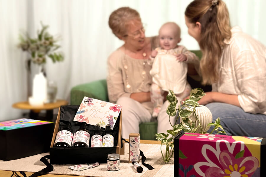 Mother's Day Gift Guide. Grandmother and mum playing with baby on couch in the background. Relax and unwind hamper open on the coffee table and box of pure relaxation.