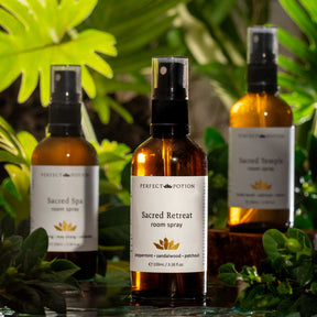 Three amber glass room spray bottles with black spray tops and white labels from Perfect Potion, including Sacred Spa, Sacred Retreat, and Sacred Temple, styled with green tropical leaves.