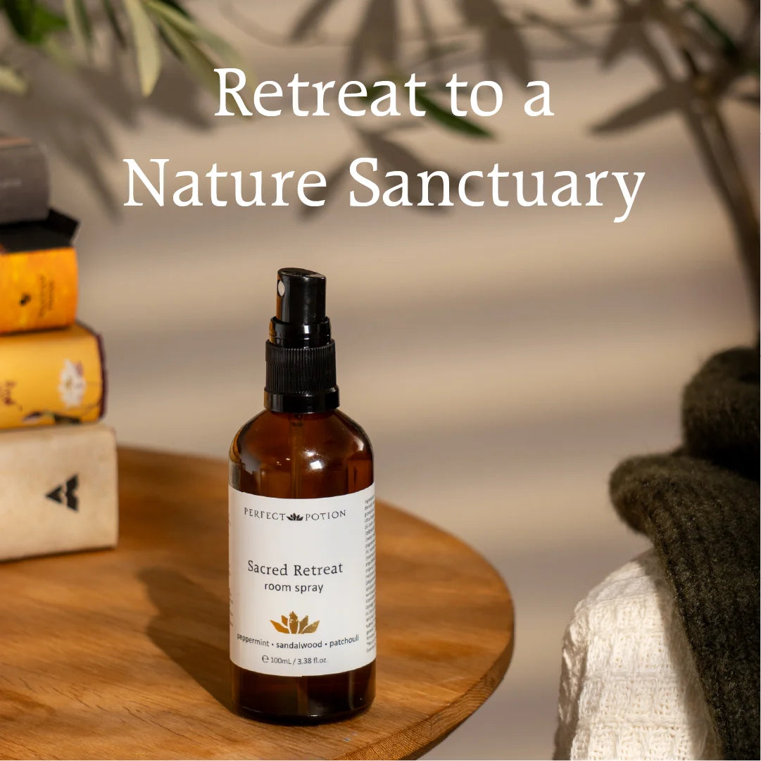 A bottle of Perfect Potion Sacred Retreat room spray stands on a round wooden table in a softly styled indoor setting with books, shadows, and a folded textile in the background. Large text at the top reads 'Retreat to a Nature Sanctuary'.
