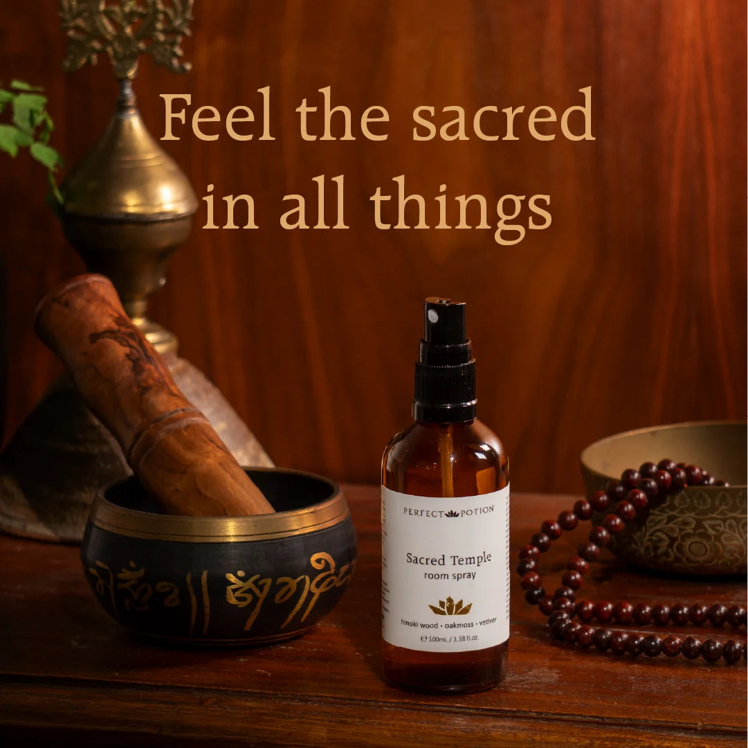 A bottle of Perfect Potions Sacred Temple Holy Water Spray stands on a wooden surface, surrounded by spiritual props including a brass bowl with a wooden mallet, a bowl of dark wooden beads, and a decorative object against a warm wood-paneled background. Text on the image reads, 'Feel the sacred in all things.